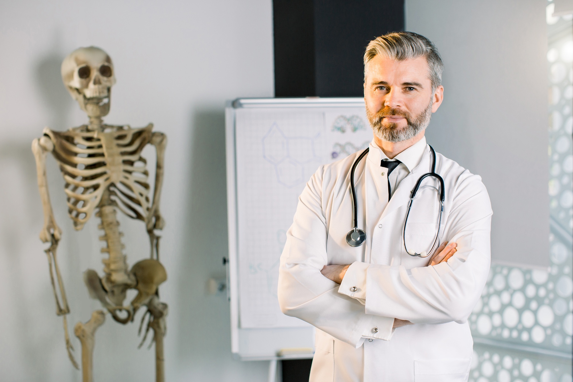 Smiling mature Caucasian man doctor posing with arms crossed in the office, wearing white coat and
